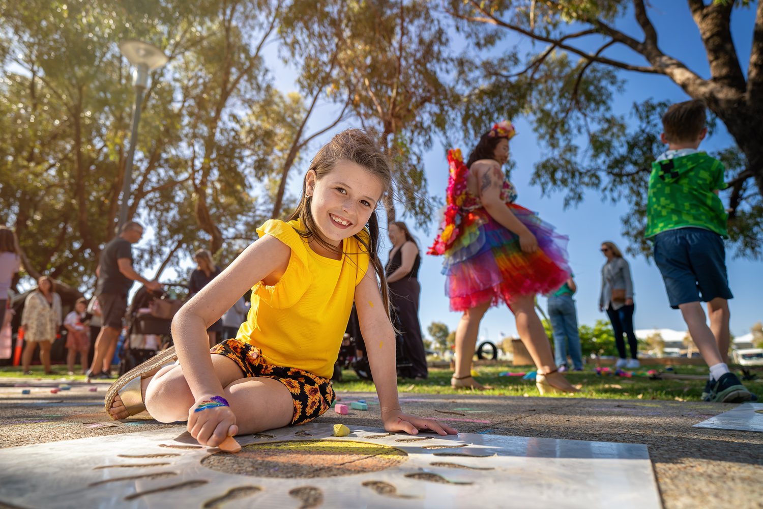 A young girl with a yellow tshirt and sunflower patterned shorts doing chalk drawing with a sun template on the pathway.